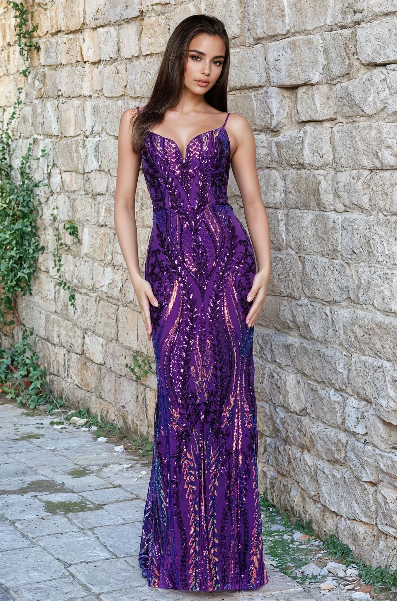 A woman poses outdoors by a stone wall in the Blondie Nites 5358BN long sequin-print prom dress, featuring intricate patterns and shimmering details in Purple Multi—an elegant evening gown available in junior sizes.